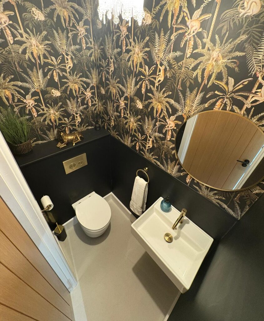 Wide shot of the entire cloakroom with a white toilet, white sink, gold-toned tap and flush buttons, and a round mirror.
