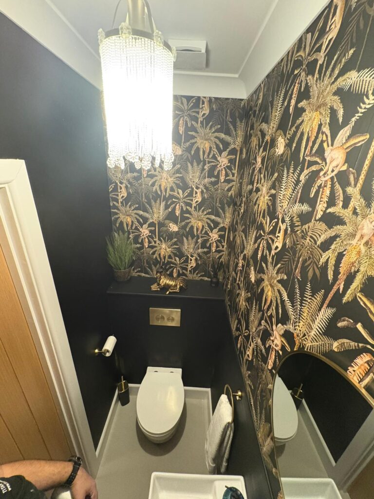 Wide shot of the cloakroom featuring a small crystal chandelier, white toilet, circular mirror and black palm-leaf walls.