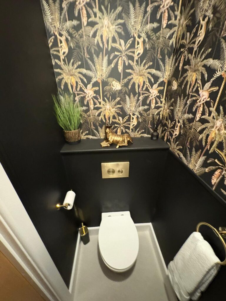 Photo of a bathroom, in the image is a standalone toilet, black wall with palm leaves.